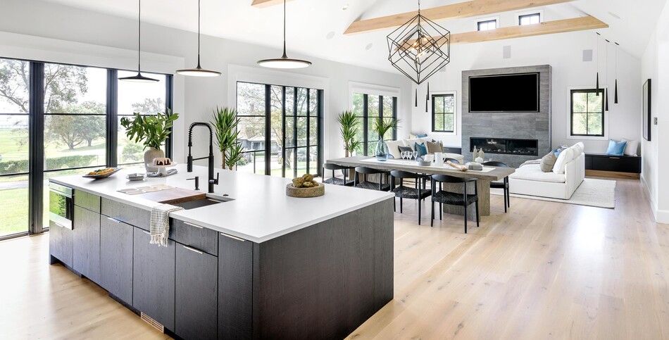 Open-concept living space with vaulted white ceiling, exposed wooden beams, a Fresh Concrete quartz kitchen island, and a dining area with a stone fireplace wall.