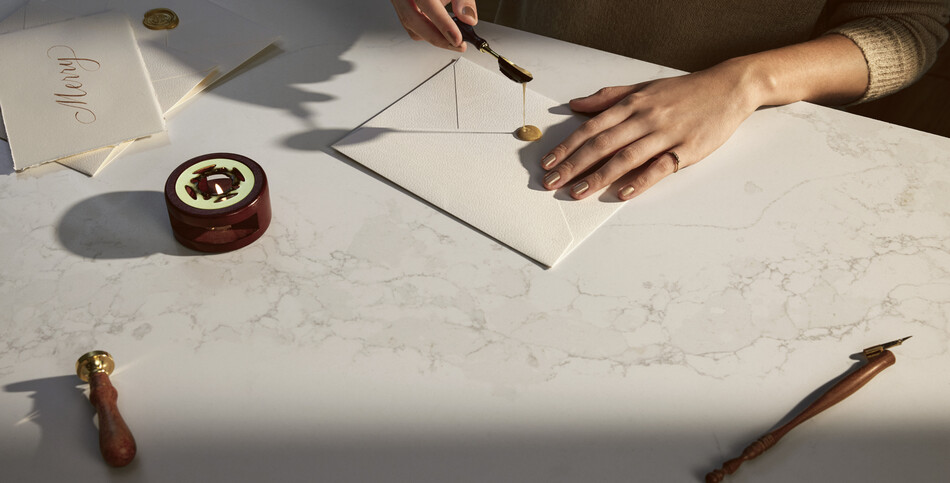Hands sealing an envelope with wax on a bright white Calacatta Stillstorm quartz countertop featuring subtle whites and pale gray veins.