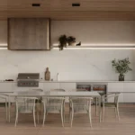 Modern outdoor kitchen with dining table, white chairs, grill, plants, and wooden ceiling.
