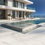 Modern luxury house with glass railings, infinity pool, loungers, and ocean view under clear sky.