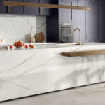 Modern kitchen with marble island, wooden stools, purple cabinets, built-in oven, and gold faucet.